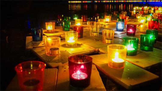 Mehrere brennende Kerzen in bunten Gläsern auf einem Holzschiffchen im Wasser bei Nacht