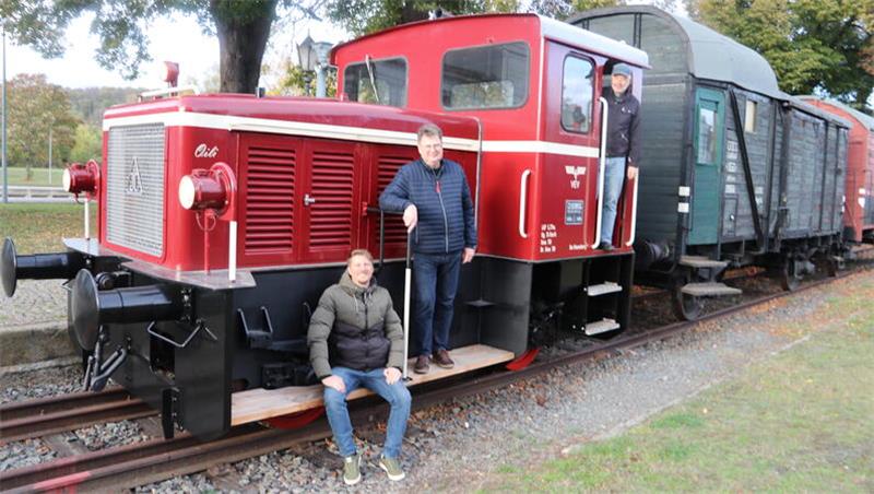 Drei Personen an einer roten Diesellok mit angehängtem Güterwagen auf Gleisen im Freien.