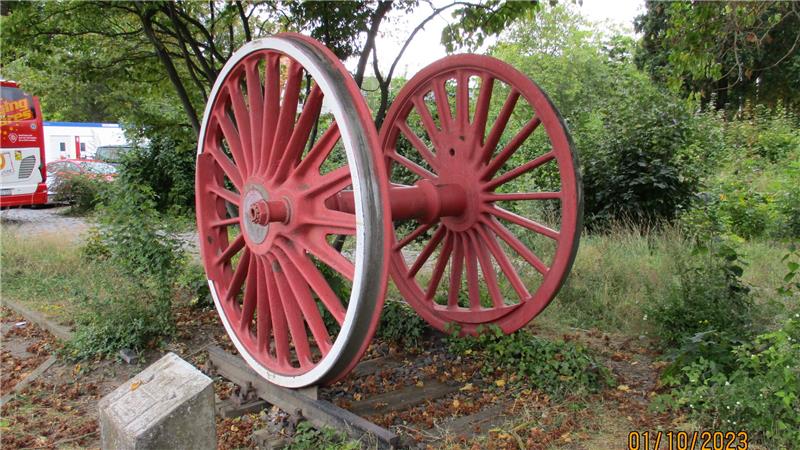 Zwei große rote Eisenbahnräder mit Speichen stehen aufrecht im Freien auf einem Gleisabschnitt neben einem Stein mit nicht lesbarer Inschrift.