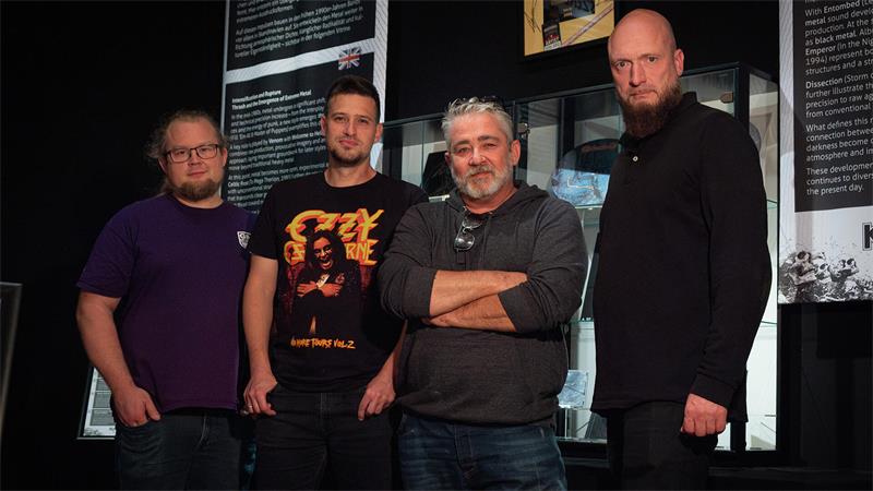Vier Männer in schwarzen Klamotten stehen nebeneinander, eine hat die Arme verschränkt.
