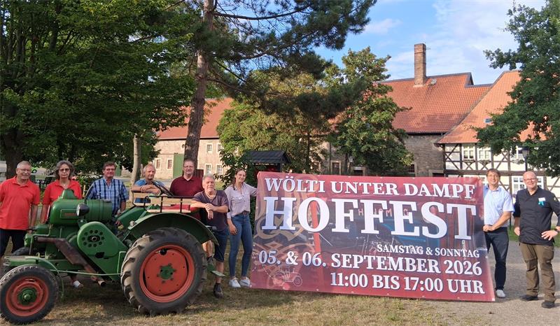 Gruppe von Menschen steht neben einem alten grünen Traktor vor einem Fachwerkhaus und hält ein Banner mit der Aufschrift 'Wölti unter Dampf Hoffest 05. & 06. September 2026 Samstag & Sonntag 11:00 bis 17:00 Uhr'