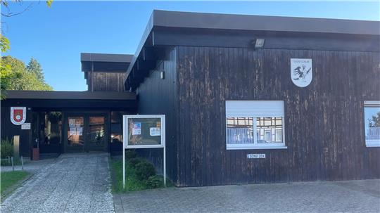 Eingangsbereich eines Gebäudes mit dunkler Holzfassade, zwei Wappen an der Fassade und einem Schild mit der Aufschrift 'SPORTHALLE'.