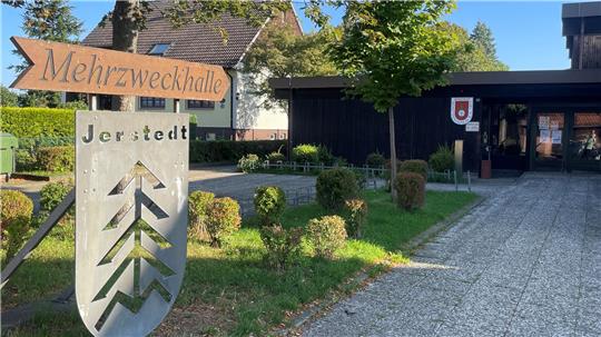 Holzschild mit der Aufschrift 'Mehrzweckhalle' neben einem Schild mit stilisierten Tannenbäumen und dem Schriftzug 'Jerstedt' vor einem Gebäude mit Eingangsbereich und Bäumen
