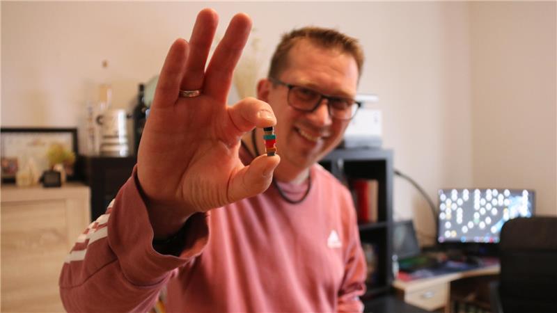 Person in rotem Pullover zeigt mit der rechten Hand ein kleines rundes Objekt zwischen Daumen und Zeigefinger.