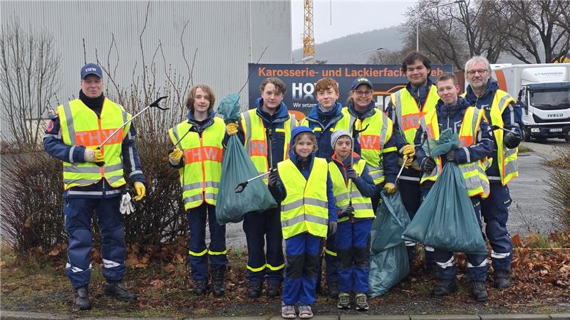 Gruppe von Menschen in gelben Warnwesten mit Müllzangen und Müllsäcken steht auf Gehweg vor Industriegebäude und Büschen.