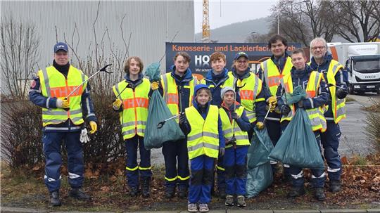 Gruppe von Menschen in gelben Warnwesten mit Müllzangen und Müllsäcken steht auf Gehweg vor Industriegebäude und Büschen.