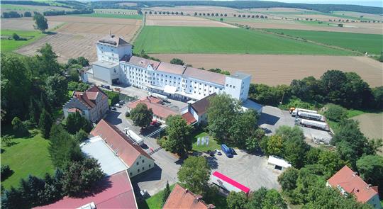 Luftaufnahme eines landwirtschaftlichen Betriebs mit mehreren Gebäuden, Fahrzeugen und Feldern im Hintergrund