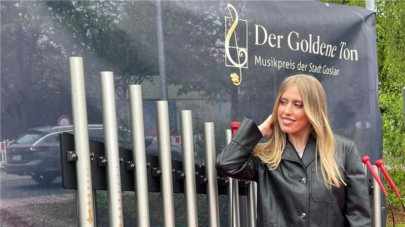 Das Foto zeigt die Musikerin Lea in einer schwarzen Jacke vor einer Klanginstallation in Hahnenklee. Ein dunkles Transparent im Hintergrund zeigt oben rechts ein Logo mit Notenschlüssel und dem Schriftzug: "Der Goldene Ton. Musikpreis der Stadt Goslar".