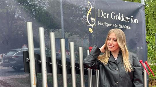 Das Foto zeigt die Musikerin Lea in einer schwarzen Jacke vor einer Klanginstallation in Hahnenklee. Ein dunkles Transparent im Hintergrund zeigt oben rechts ein Logo mit Notenschlüssel und dem Schriftzug: "Der Goldene Ton. Musikpreis der Stadt Goslar".
