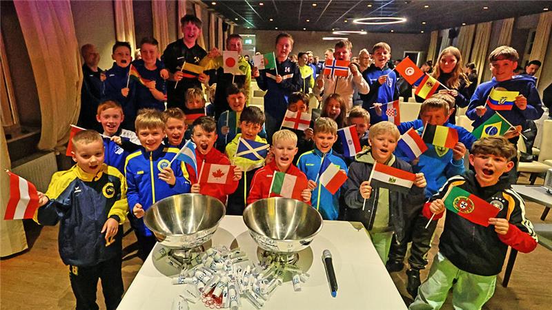 Gruppe von Kindern in Sportkleidung hält verschiedene Länderflaggen in einem Raum mit zwei großen Metallbehältern und einem Mikrofon auf einem Tisch im Vordergrund