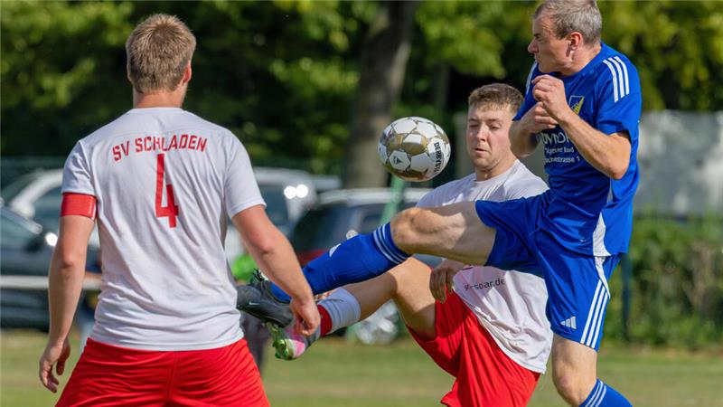Drei Fußballspieler kämpfen um den Ball, zwei in weißen Trikots mit roter Schrift und einer in blauem Trikot mit erhobenem Bein.