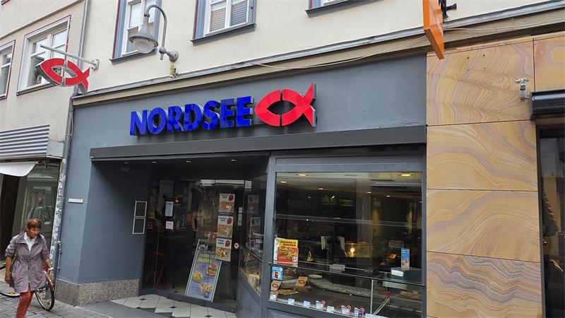 Fassade eines Nordsee-Restaurants mit blauem Schriftzug und rotem Fischsymbol über dem Eingang in einer Fußgängerzone. 