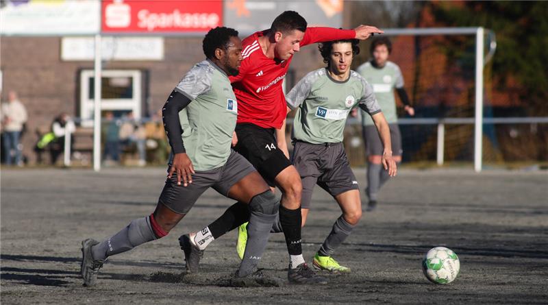 Drei Fußballspieler kämpfen um den Ball auf einem Spielfeld mit Tor im Hintergrund.