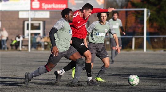 Drei Fußballspieler kämpfen um den Ball auf einem Spielfeld mit Tor im Hintergrund.