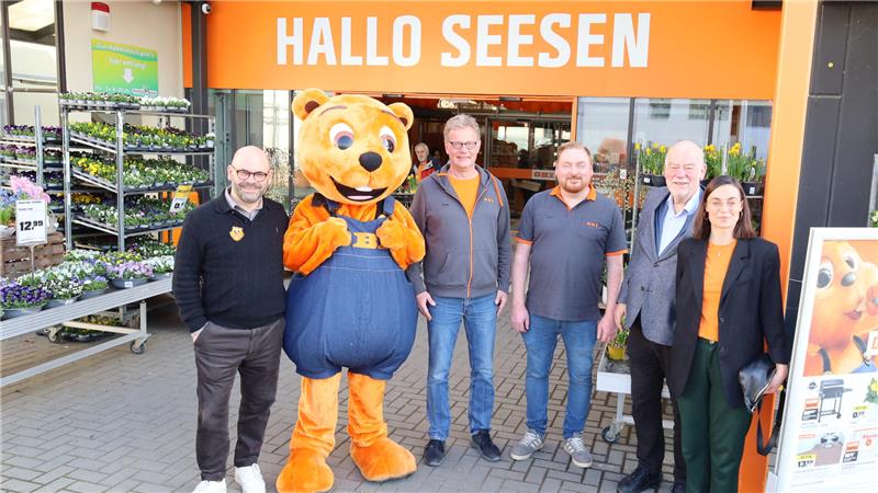 Fünf Personen und eine Biberfigur in blauer Latzhose stehen vor einem orangefarbenen Eingang mit der Aufschrift 'HALLO SEESEN'