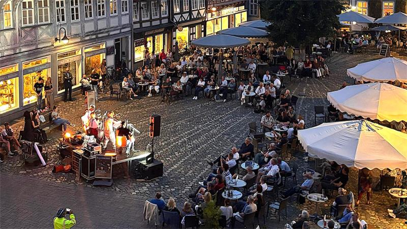 Abendliche Straßenszene mit Live-Musik auf einem gepflasterten Platz vor Fachwerkhäusern und zahlreichen Sitzgruppen unter großen Sonnenschirmen.
