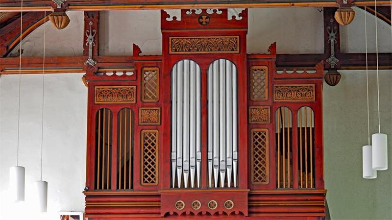 Rote Orgel mit silbernen Pfeifen in einer Kirche mit sichtbaren Holzbalken und weißen Hängelampen