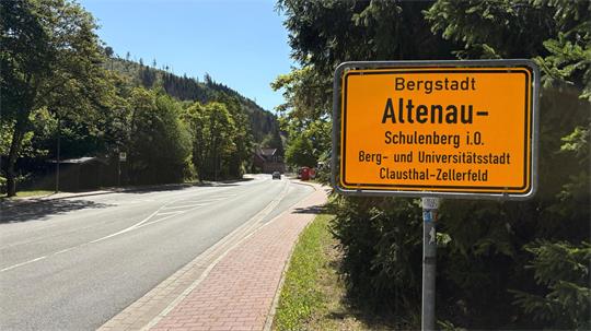 Ortseingangsschild mit der Aufschrift 'Bergstadt Altenau-Schulenberg i.O. Berg- und Universitätsstadt Clausthal-Zellerfeld' an einer Landstraße mit Bäumen und Gehweg.