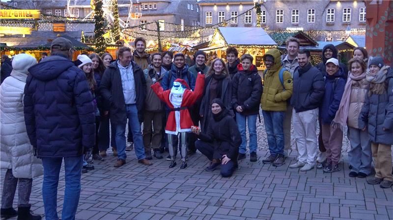 Gruppe von Menschen steht auf gepflastertem Platz vor weihnachtlich geschmückten Holzhütten, eine Person in Weihnachtsmannkostüm hebt die Arme.