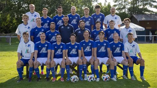 Mannschaftsbild einer Fußballmannschaft mit Spielern und Trainern in blauen Trikots und weißen Poloshirts auf einer Wiese vor Bäumen