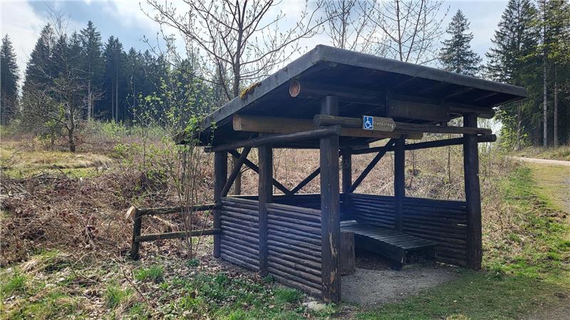 Holzunterstand mit Sitzbank und Dach in einer Waldlichtung neben einem Weg
