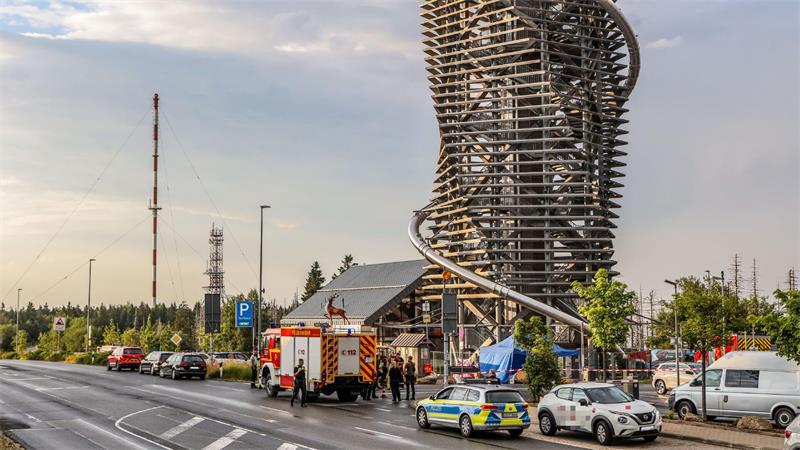 Straße mit Einsatzfahrzeugen und Personen vor einem hohen, spiralförmig umwickelten Aussichtsturm aus Holz.