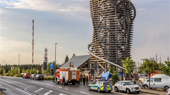 Straße mit Einsatzfahrzeugen und Personen vor einem hohen, spiralförmig umwickelten Aussichtsturm aus Holz.