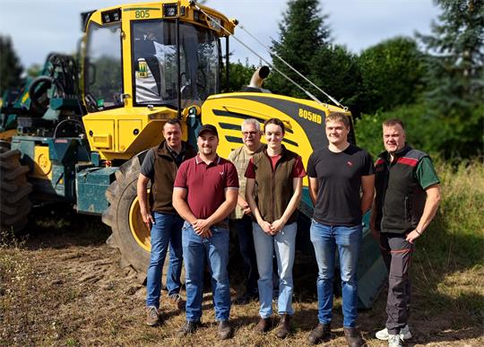 Sechs Personen stehen vor einem gelben Baumaschinenfahrzeug auf einem Feld mit Bäumen im Hintergrund