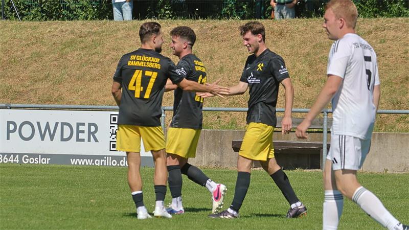 Drei Fußballspieler in schwarzen Trikots mit gelben Hosen feiern auf dem Spielfeld, ein Spieler in weißem Trikot geht vorbei.