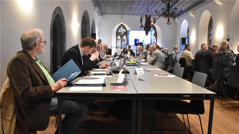 Menschen sitzen an einem langen Tisch in einem historischen Raum mit hohen Fenstern und Kronleuchtern und arbeiten oder diskutieren