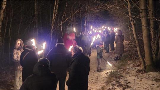 Man sieht mehrere Wanderer mit einer Fackel, die über einen schneebedeckten Weg durch den Wald gehen.