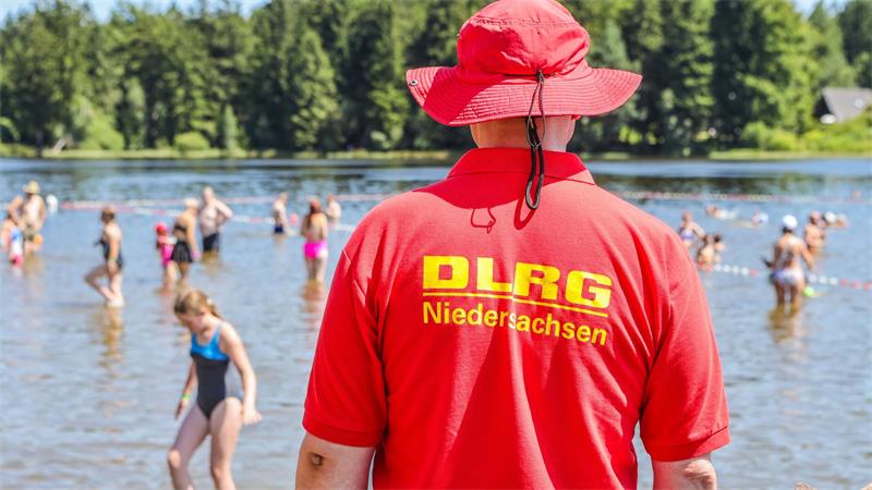Rettungsschwimmer in rotem T-Shirt mit Aufschrift 'DLRG Niedersachsen' steht am Ufer eines Sees, im Wasser spielen mehrere Kinder.