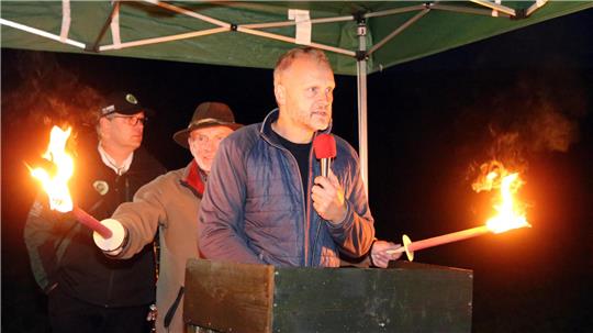 Alexander Ehrenberg steht mit Mikro am Rednerpult. Im Hintergrund hält jemand Fackeln.