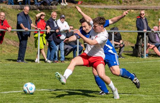 Zwei Fußballspieler in Aktion auf dem Spielfeld, einer in weißem Trikot mit roten Hosen, der andere in blauem Trikot, im Hintergrund Zuschauer auf einer Tribüne