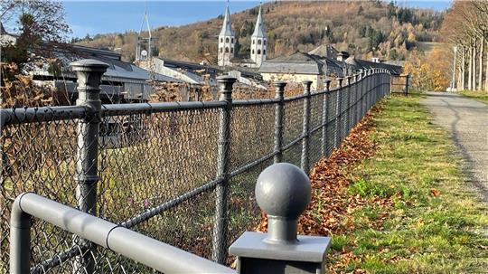 Zu sehen ist ein Maschendrahtzaun am Wegesrand mit zwei Kirchtürmen und Bergen im Hintergrund.