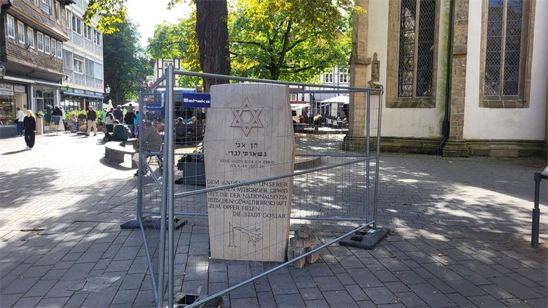 Steinerner Gedenkstein mit Davidstern und hebräischer sowie deutscher Inschrift auf einem gepflasterten Platz, umgeben von einem Bauzaun, im Hintergrund Menschen und historische Gebäude.