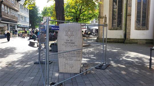 Steinerner Gedenkstein mit Davidstern und hebräischer sowie deutscher Inschrift auf einem gepflasterten Platz, umgeben von einem Bauzaun, im Hintergrund Menschen und historische Gebäude.
