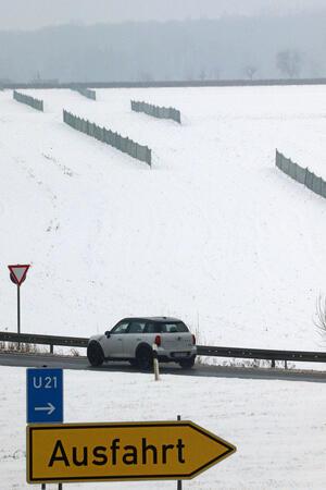 Schneefangzäune auf einem Feld, im Vordergrund kennzeichnet ein Schild eine Abfahrt, dahinter fährt ein Auto.