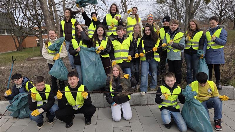 Gruppe von Menschen mit Warnwesten und Handschuhen hält Müllsäcke und Greifzangen im Freien vor kahlen Bäumen.
