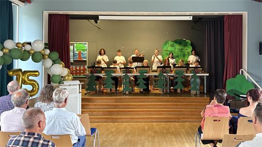 Schülerinnen und Schüler spielen auf einer Bühne vor Publikum in einem Saal mit Holzdecke und blauen Vorhängen.
