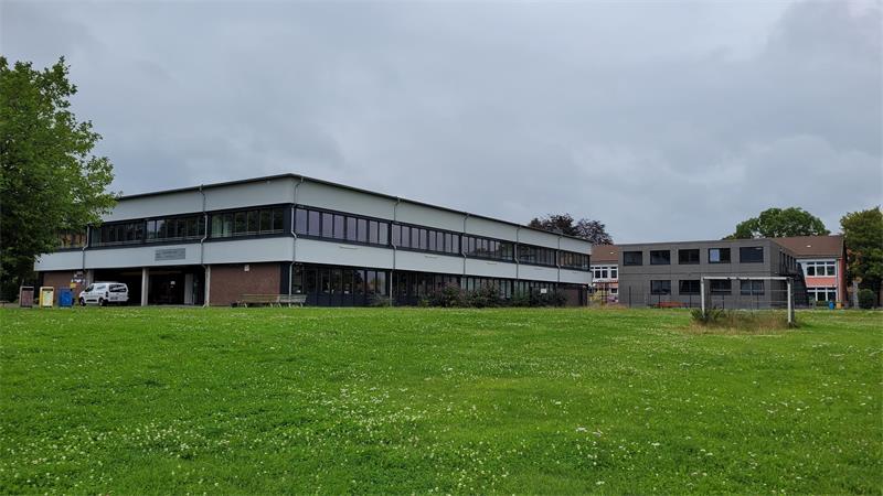 Das Bild zeigt eine Wiese mit einem Bolzplatz, daginter ein großes Schulgebäude.