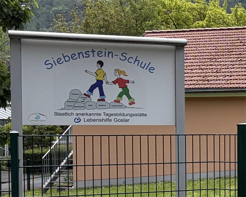 Schild der Siebenstein-Schule mit zwei Kindern, die über beschriftete Steine laufen, vor einem Gebäude mit rotem Ziegeldach und einem Metallzaun im Vordergrund.