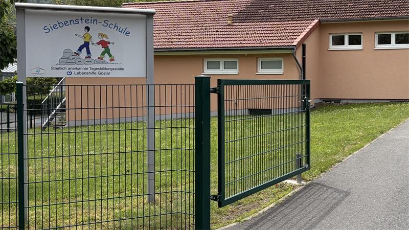 Eingangstor mit grünem Metallzaun vor einem hellen Schulgebäude und einem Schild mit der Aufschrift 'Siebenstein-Schule'