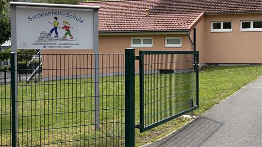 Eingangstor mit grünem Metallzaun vor einem hellen Schulgebäude und einem Schild mit der Aufschrift 'Siebenstein-Schule'