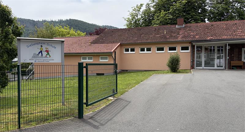 Eingang eines eingeschossigen Gebäudes mit rotem Ziegeldach, grünem Metallzaun und einem Schild mit bunter Zeichnung neben dem Tor