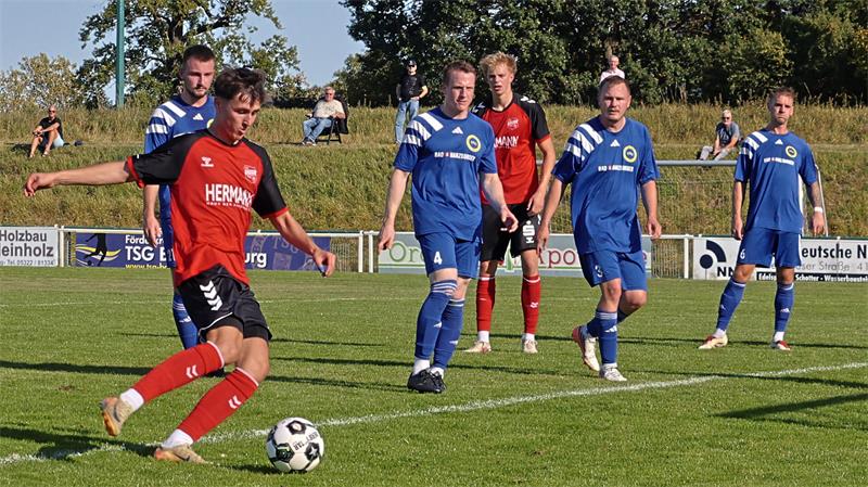 Fußballspieler in blauen und roten Trikots auf dem Spielfeld, ein Spieler in rotem Trikot schießt den Ball