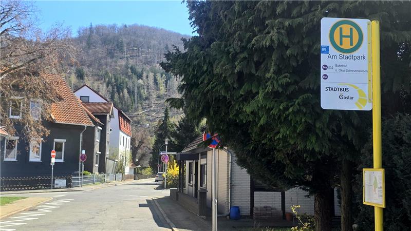 Straße mit Bushaltestelle und Haltestellenschild vor Häusern und bewaldetem Hügel im Hintergrund.
