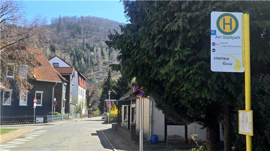 Straße mit Bushaltestelle und Haltestellenschild vor Häusern und bewaldetem Hügel im Hintergrund.