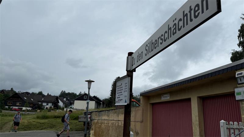 Das Foto zeigt ein Straßenschild im Vordergrund und zwei Wanderer vor einem Wohngebiet im Hintergrund.