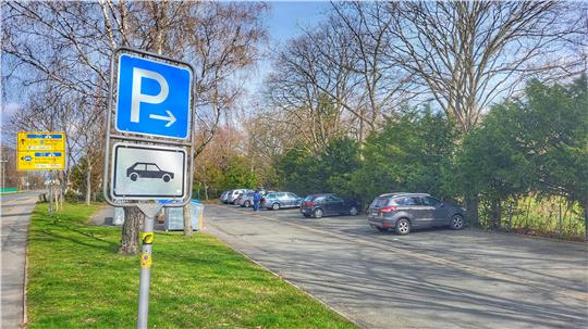 Parkplatzschild mit Pfeil nach rechts vor einem Parkplatz mit mehreren geparkten Autos und Bäumen im Hintergrund